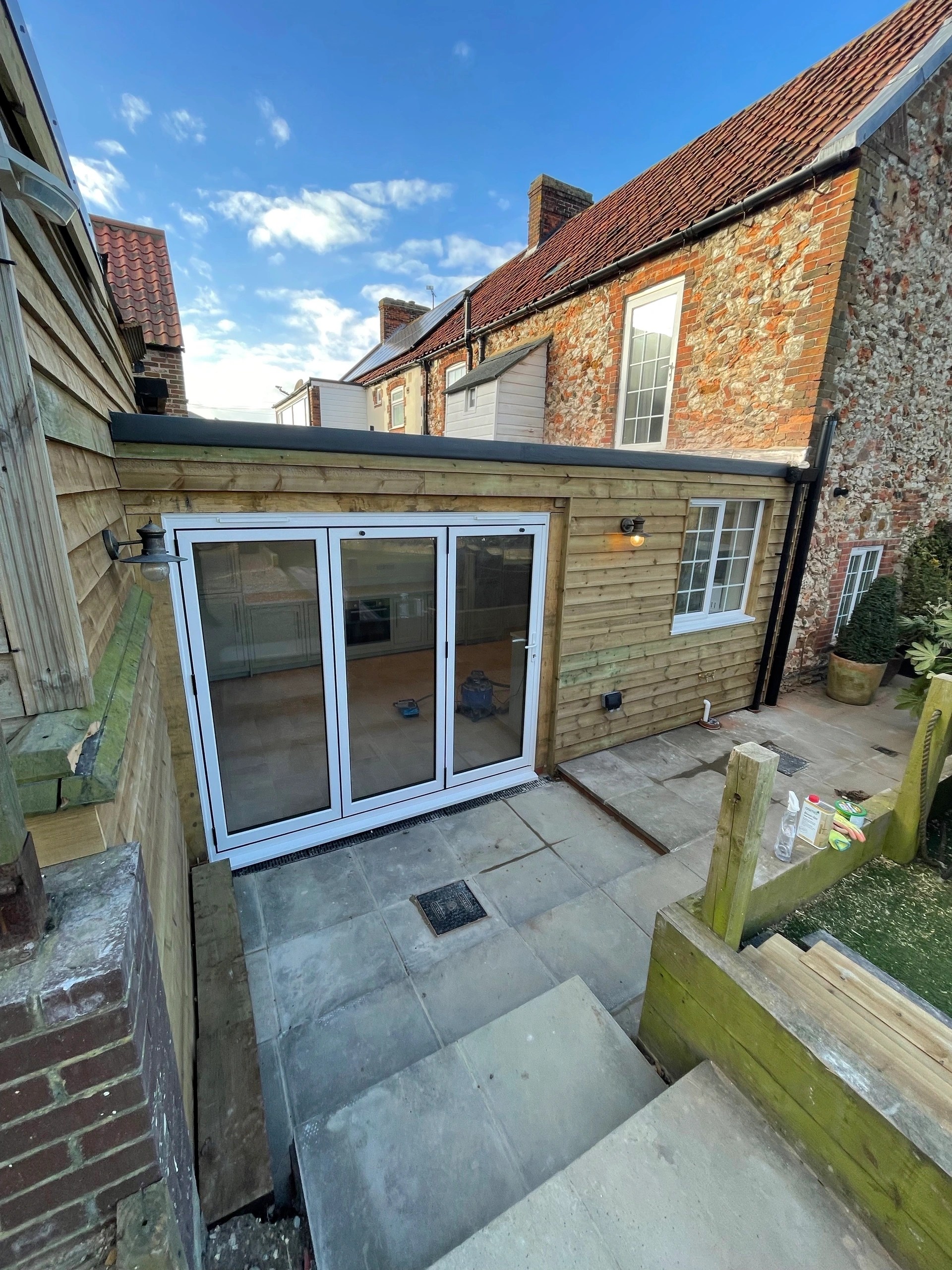 Timber-clad extension to a flint and brick cottage, with white bi-fold doors onto a paved courtyard